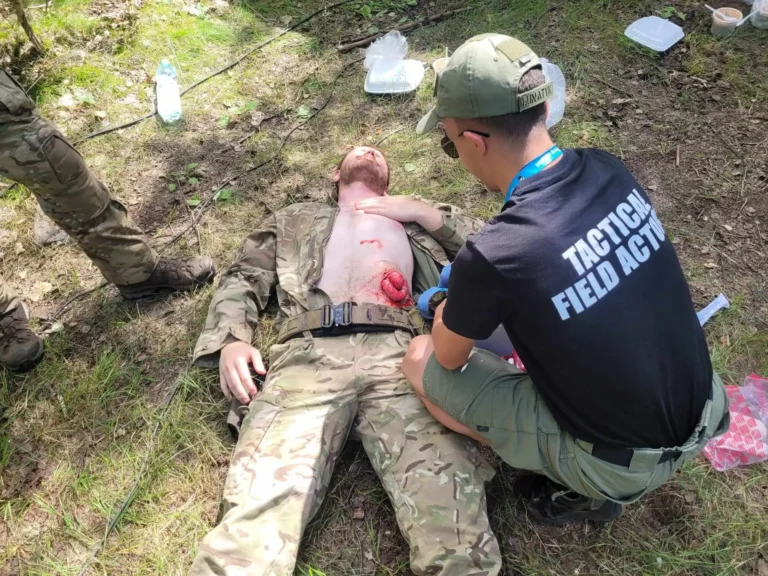 Instruktor w koszulce „Tactical Field Actors” udziela pomocy osobie z pozorowaną raną podczas treningu ratownictwa taktycznego na trawie.