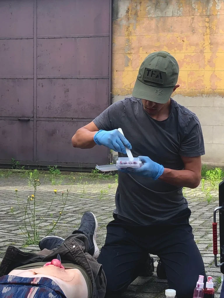 Mężczyzna w czapce z napisem TFA przygotowujący masę do pozoracji ran (moulage) nad poszkodowanym z raną jamy brzusznej.