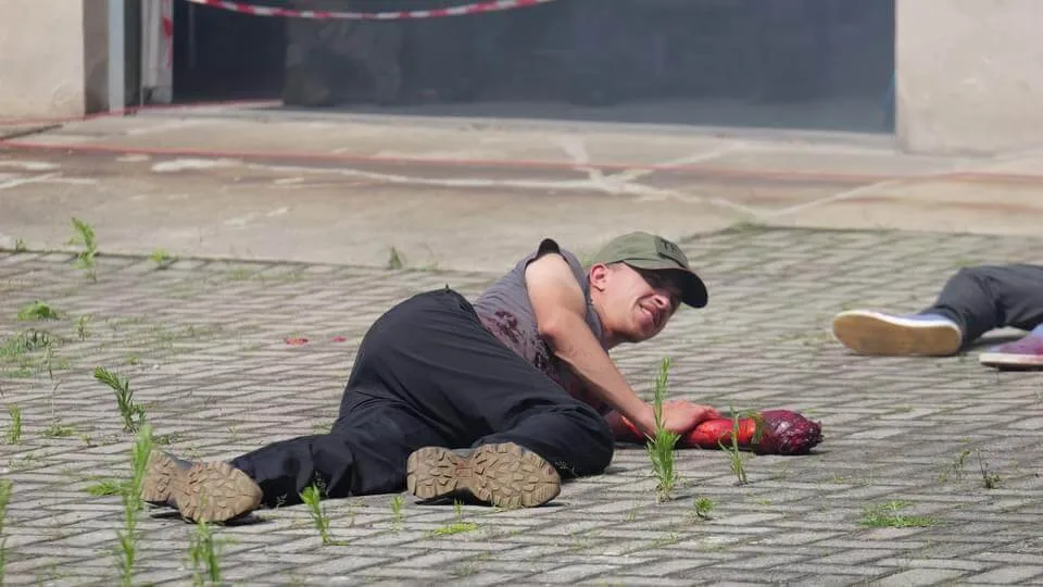Pozorant leżący na kostce brukowej podczas ćwiczeń Tactical Public Rescue 2024, z realistyczną pozoracją ciężkiej rany dłoni i krwawieniem na przedramieniu.